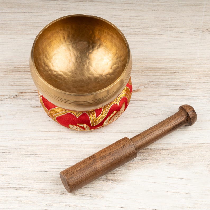 May include: A gold-coloured metal singing bowl with a hammered texture, resting on a circular red fabric cushion featuring gold embroidered patterns. A cylindrical wooden mallet with a smooth finish is positioned next to the bowl.