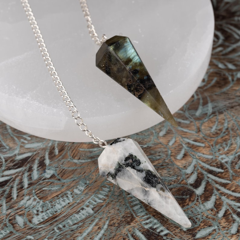 May include: Two gemstone pendulums with silver chains. One pendulum is a dark green and brown color, the other is white with black markings. The pendulums are on a white surface and a wooden surface.