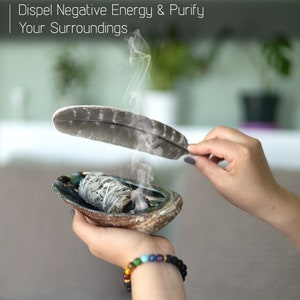 Pu&ograve; includere: Una mano tiene una conchiglia con un fascio di salvia fumante al suo interno. Una piuma bianca &egrave; tenuta sopra la conchiglia, con fumo che sale dalla salvia. Il testo "Dispel Negative Energy & Purify Your Surroundings" &egrave; in cima all'immagine.