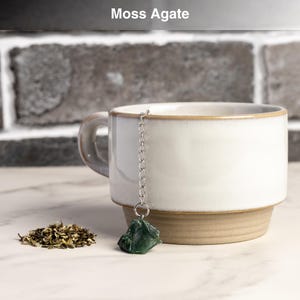May include: A white ceramic mug with a brown base and rim, holding a silver chain with a green moss agate tea infuser. A small pile of loose tea leaves sits beside the mug. The text "Moss Agate" is above the mug.
