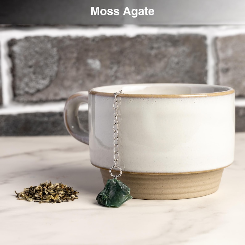 May include: A white ceramic mug with a brown base and rim, holding a silver chain with a green moss agate tea infuser. A small pile of loose tea leaves sits beside the mug. The text "Moss Agate" is above the mug.