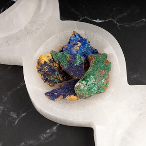 May include: A collection of blue, green, and brown mineral specimens in a white selenite bowl. The bowl is shaped like a crescent moon.