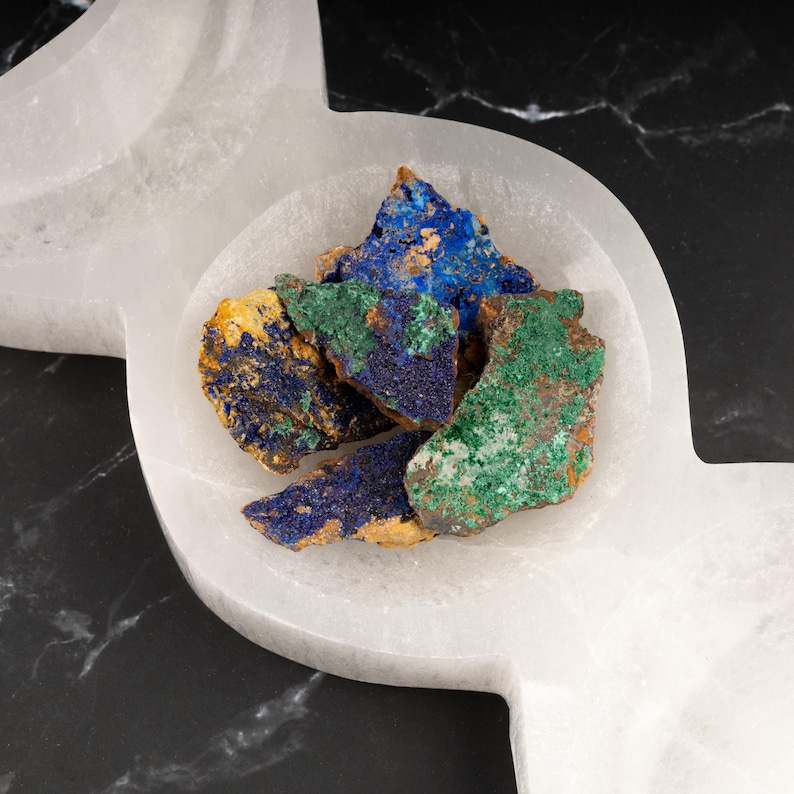 May include: A collection of blue, green, and brown mineral specimens in a white selenite bowl. The bowl is shaped like a crescent moon.
