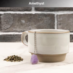 May include: A white ceramic mug with a brown base and rim, paired with a purple amethyst tea infuser. A small pile of loose tea leaves sits beside the mug. The word "Amethyst" is at the top of the image.