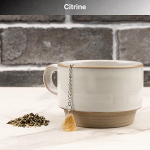 May include: A white ceramic mug with a brown base and rim, a silver chain, and a yellow citrine crystal tea infuser. A small pile of green tea leaves sits next to the mug. The word "Citrine" is at the top.
