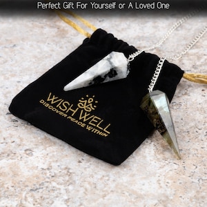 May include: Two crystal pendulums with silver chains rest on a black velvet bag. One pendulum is white with black markings, the other is gray and green. The bag has gold text that reads "WISHWELL DISCOVER PEACE WITHIN."