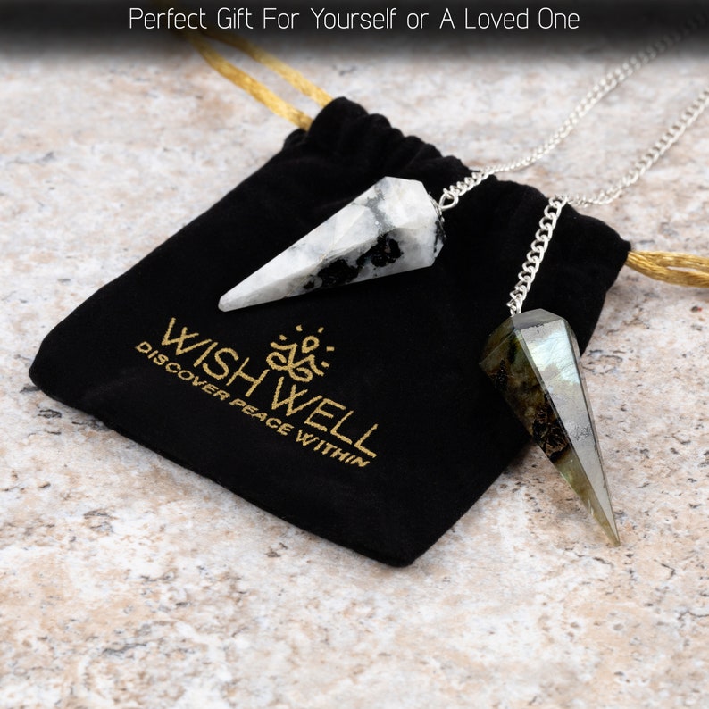 May include: Two crystal pendulums with silver chains rest on a black velvet bag. One pendulum is white with black markings, the other is gray and green. The bag has gold text that reads "WISHWELL DISCOVER PEACE WITHIN."