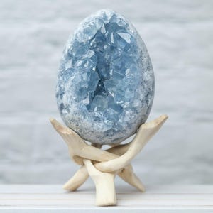 May include: A large, oval-shaped celestite crystal geode, showcasing a captivating light blue hue and crystalline interior. It is displayed on a natural wooden stand with a three-legged design, creating a striking contrast.