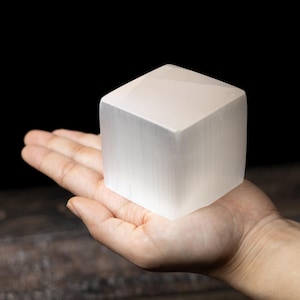 May include: A translucent, white selenite cube is held in an open hand. The cube has a smooth, polished surface and a geometric shape. The background is dark, highlighting the cube's crystalline structure. The cube is a decorative item.