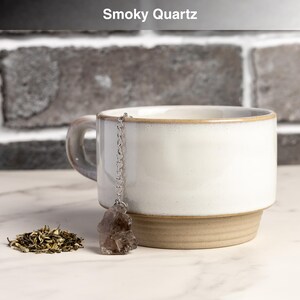 May include: A white ceramic mug with a tan base and rim, holding a smoky quartz tea infuser. The infuser is attached to a silver chain. A small pile of loose tea leaves sits beside the mug. The text "Smoky Quartz" is at the top.