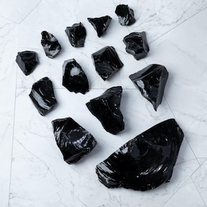 May include: A collection of polished, black obsidian stones of varying sizes and shapes. The stones are arranged on a white marble-patterned surface, reflecting light and showcasing their glossy texture. The obsidian pieces are a deep, opaque black.
