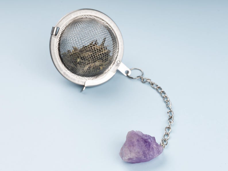 May include: A stainless steel tea infuser with a chain and a purple amethyst stone. The infuser is filled with loose tea leaves. The tea infuser is round and has a mesh design.
