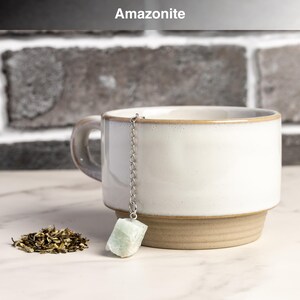 May include: A white ceramic mug with a light brown base and rim, holding a tea infuser with a light green amazonite stone. A small pile of loose tea leaves sits beside the mug. The word "Amazonite" is at the top.