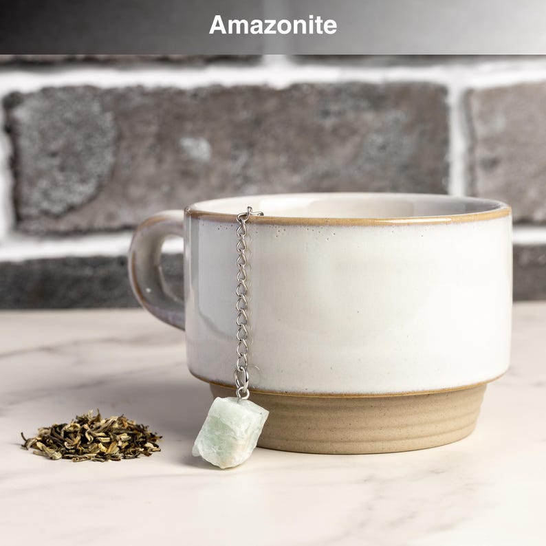 May include: A white ceramic mug with a light brown base and rim, holding a tea infuser with a light green amazonite stone. A small pile of loose tea leaves sits beside the mug. The word "Amazonite" is at the top.
