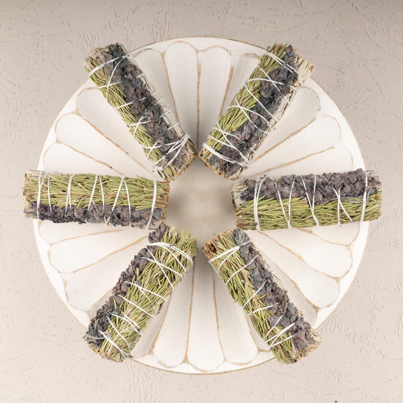 May include: Six bundles of dried sage and lavender smudge sticks arranged in a circle on a white wooden plate. The smudge sticks are tied with twine and have a natural, earthy look.
