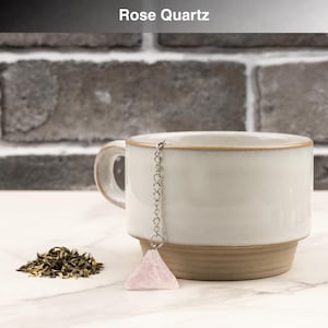 May include: A white ceramic mug with a brown base and rim, paired with a rose quartz tea infuser. The infuser is a pink, triangular crystal suspended by a silver chain. A small pile of loose tea leaves sits beside the mug. The text "Rose Quartz" is at the top.
