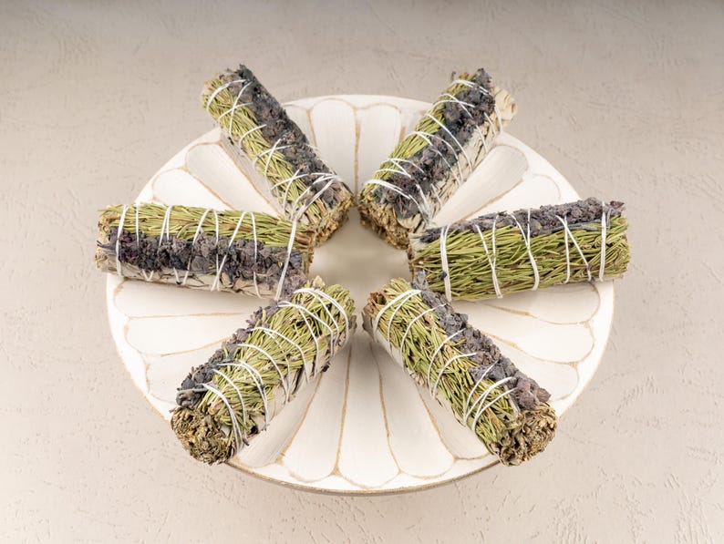 May include: Six bundles of dried herbs, including sage and lavender, are arranged in a starburst pattern on a white ceramic plate. The bundles are tied with twine and are ready to be used for smudging.