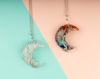 May include: Two crescent moon pendants on silver chains. One is clear with silver wire detailing, the other is multi-colored with red, orange, blue, and purple stones. The background is split between teal and peach.