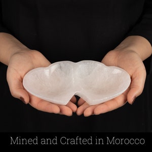 May include: A white selenite heart-shaped dish with two compartments. The dish is being held in the hands of a person.