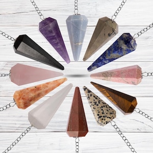 May include: A set of 12 crystal pendulums in various colors and shapes, including amethyst, rose quartz, and tiger's eye. The pendulums are hanging from silver chains and are arranged in a circle on a white wooden background.