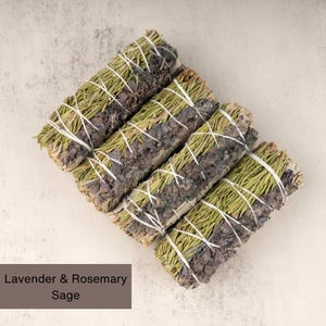 May include: Four bundles of dried sage smudge sticks tied with twine. The sage is a blend of lavender and rosemary. The sticks are arranged in a stack on a light beige surface.  Lavender & Rosemary Sage