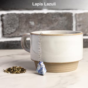 May include: A white ceramic mug with a tan base and rim, holding a tea infuser. The infuser features a chain and a blue and white Lapis Lazuli stone. The text "Lapis Lazuli" is at the top. A small pile of loose tea leaves sits nearby.