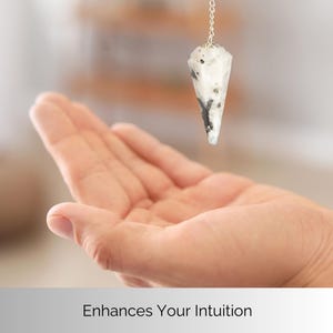 May include: A white and black crystal pendulum suspended from a silver chain. The pendulum is cone-shaped and hangs above an open hand. The text "Enhances Your Intuition" is at the bottom of the image.