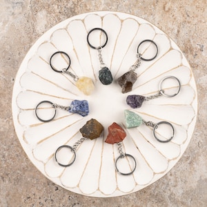 May include: A collection of keychains featuring various colorful raw gemstones. Each keychain has a silver-tone chain and a black key ring. The gemstones include yellow, green, brown, purple, blue, and red stones. The keychains are arranged on a white, scalloped, wooden tray.
