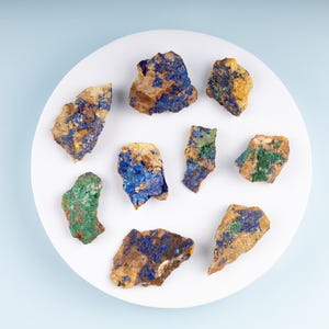 May include: A collection of nine small, rough, irregularly shaped stones with blue, green, and yellow mineral deposits on a white plate.