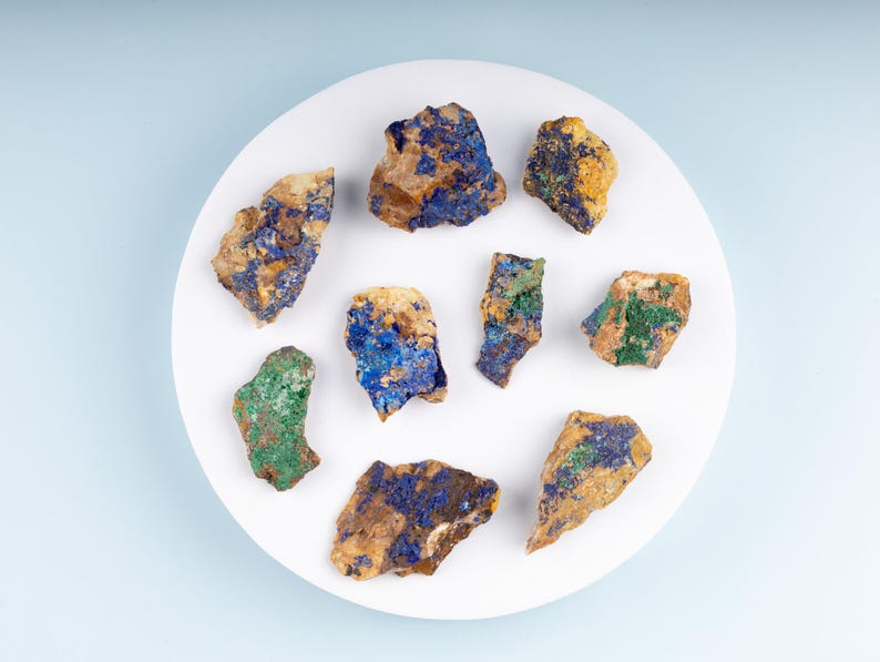 May include: A collection of nine small, rough, irregularly shaped stones with blue, green, and yellow mineral deposits on a white plate.