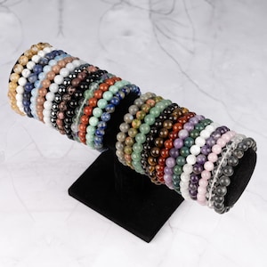 May include: A collection of colorful gemstone bracelets stacked on a black display stand. The bracelets are made with various types of stones, including amethyst, rose quartz, tiger's eye, and clear quartz.