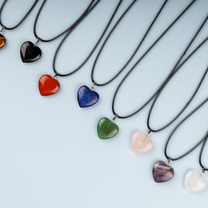 May include: A collection of heart-shaped gemstone pendants on black cords. The pendants are in various colors, including brown, black, red, blue, green, pink, purple, and white. The necklaces are displayed against a light blue background.