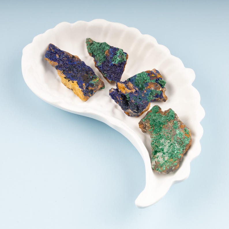May include: Four pieces of malachite and azurite rough stone on a white ceramic dish. The stones are green, blue, and gold in color.