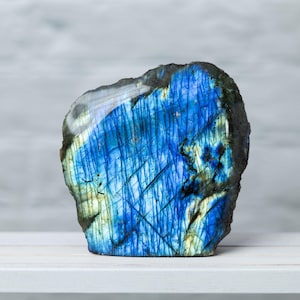 May include: A polished labradorite gemstone with a striking display of blue and green iridescence. The stone has a rough, natural edge and sits on a white surface. The gemstone's surface is marked with linear patterns.