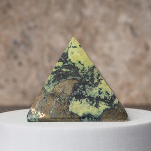 May include: A pyramid-shaped decorative stone with a mix of green, black, and brown colors. The pyramid is displayed on a white surface, showcasing its geometric form and natural patterns. The stone's texture and color variations create a unique aesthetic.