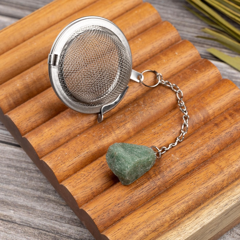 May include: A stainless steel tea infuser with a chain and a green stone charm rests on a wooden tray. The infuser has a mesh basket and a hinged lid. The wooden tray has a ridged design.