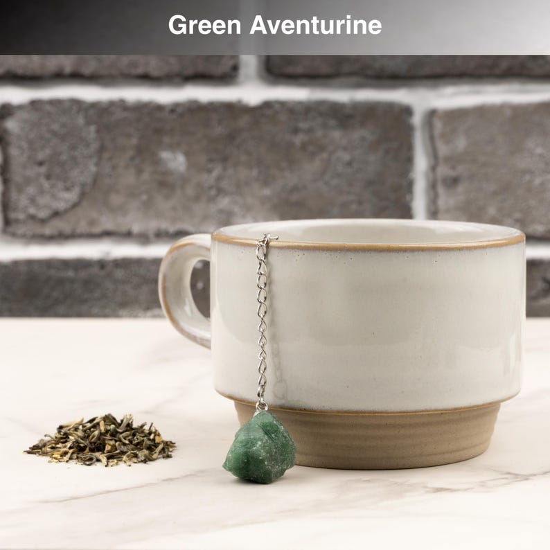 May include: A white ceramic mug with a brown base and rim, paired with a green aventurine tea infuser. The infuser features a chain and a raw, green aventurine stone. Loose tea leaves are visible beside the mug. The text "Green Aventurine" is at the top.