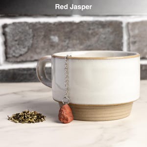 May include: A white ceramic mug with a brown base and rim, paired with a red jasper tea infuser. The infuser hangs from a silver chain. A small pile of loose tea leaves sits beside the mug. The text "Red Jasper" is at the top.