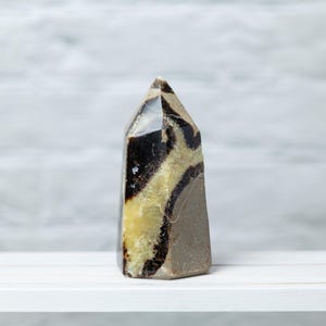 May include: A polished, rectangular crystal tower with a pointed top. The stone features a mix of colors, including black, tan, and light brown, with a marbled pattern. The crystal is displayed on a white surface against a light gray background.