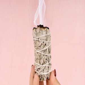 Pu&ograve; includere: Un bastoncino di salvia bianca sta bruciando, rilasciando fumo su uno sfondo rosa. La salvia &egrave; legata con uno spago bianco ed &egrave; tenuta nella mano di una persona.