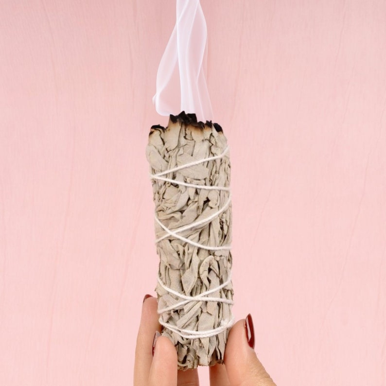 Pu&ograve; includere: Un bastoncino di salvia bianca sta bruciando, rilasciando fumo su uno sfondo rosa. La salvia &egrave; legata con uno spago bianco ed &egrave; tenuta nella mano di una persona.