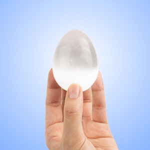 May include: A translucent, egg-shaped stone held against a gradient blue background. The stone is smooth and appears to be made of a crystalline material. The object is held in a hand, with the fingers and thumb visible.