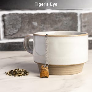 May include: A white ceramic mug with a brown base and handle, holding a tea infuser with a Tiger's Eye stone. A small pile of loose tea leaves sits beside the mug. The text "Tiger's Eye" is at the top.