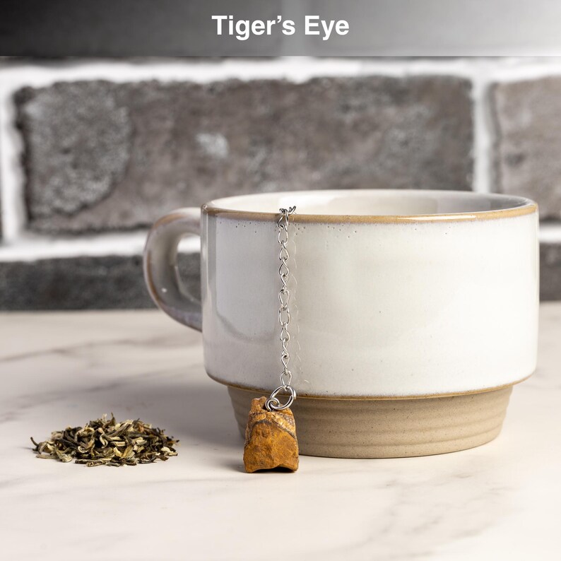 May include: A white ceramic mug with a brown base and handle, holding a tea infuser with a Tiger's Eye stone. A small pile of loose tea leaves sits beside the mug. The text "Tiger's Eye" is at the top.