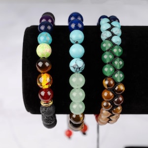 May include: Three beaded bracelets with different colored stones. The bracelets are stacked on a black display stand. The bracelets are made with natural stones in shades of blue, green, brown, and orange.
