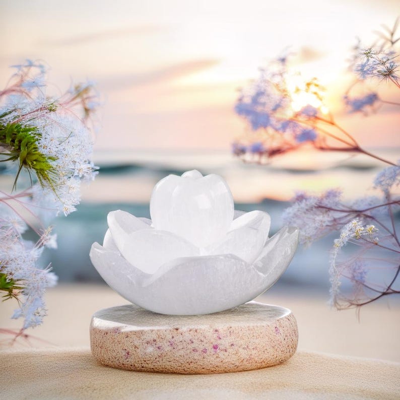 May include: A white crystal flower sculpture sits on a round, textured stone base. The sculpture is in the shape of a lotus flower with five petals. The background is a blurred image of a beach with a sunset.