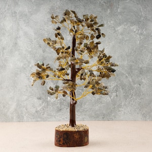 May include: A wire-wrapped crystal tree with a brown base and green and gold crystals. The tree is on a light beige surface.