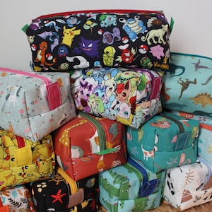 Op de afbeelding: Een stapel kleurrijke ritszakjes met verschillende cartoon- en dierenprints. De zakjes zijn gemaakt van stof en hebben een verscheidenheid aan kleuren, waaronder blauw, groen, rood, geel en zwart.