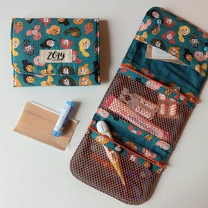 Può includere: Un organizer in tessuto color verde acqua con un motivo ripetuto di volti diversi e un'etichetta ZOLY CREATIONS. L'organizer è mostrato sia piegato che aperto, rivelando diversi scomparti in rete con cerniera, un balsamo labbra e un cerotto.
