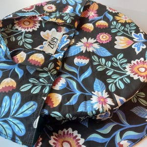 Peut inclure: Un chapeau de soleil noir avec un motif floral coloré. Le chapeau présente un motif répété de fleurs et de feuilles dans les tons de rose, bleu, jaune et blanc. L'étiquette indique "Zoly".